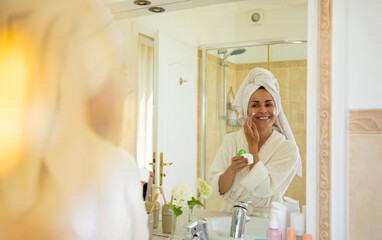A woman in a bathrobe and towel applies face cream while smiling in front of the bathroom mirror....