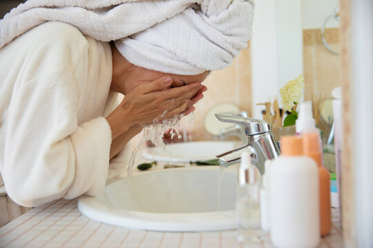 A woman in a bathrobe and towel washes her face over a bathroom sink with running water. The bright, clean setting emphasizes a refreshing skincare routine, self-care, and a morning wellness ritual.
