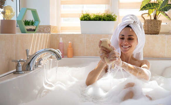 Smiling female enjoys a bubbly bath, squeezing a bath sponge under running water. Wrapped in a towel, she sits in a bright bathroom with plants and natural light, creating a peaceful self-care scene