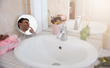 A man is seen smiling in a small round mirror while applying cream to his face. The bathroom counter features skincare products and a sink, creating a calm, self-care scene with soft natural light.