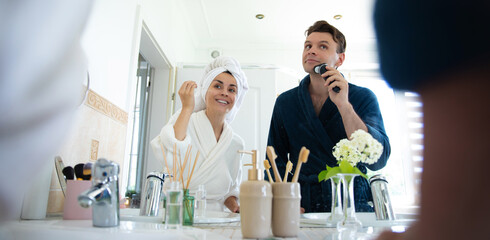 A couple in bathrobes enjoys their morning routine. The woman uses a lotion while the man shaves...