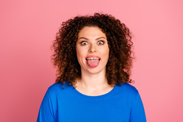 Playful young woman with curly hair sticks out tongue against vibrant pink background, wearing casual blue sweatshirt