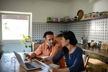 Couple managing bills with smartphone and laptop on kitchen desk at home