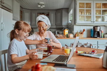 Mother and daughter having breakfast and watching laptop together in cozy kitchen