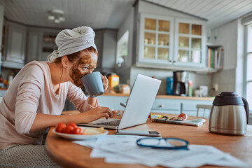 Woman with face mask working on laptop in kitchen during breakfast