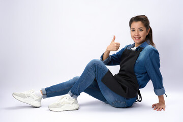 Young woman in apron showing thumb up gesture, sitting on white background, full length portrait