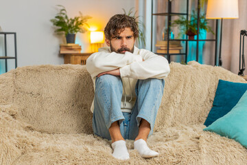 Sad frustrated young Indian man sitting at home looks pensive thinks over lonely life or problems,...