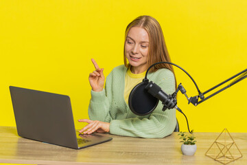 Happy excited woman blogger at table recording podcast, giving workshop, webinar. Adult girl influencer speaking into microphone and laptop webcam, making video call, discussing on yellow background.