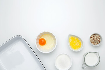 Empty white baking dish with ingredients for baking, including egg, milk, butter, sugar, and nuts on a white background. Flat lay composition for cooking preparation