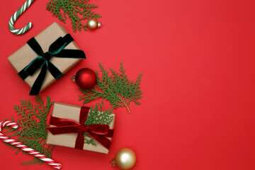 Gifts wrapped in craft paper, Christmas ornaments, thuja branches, and candy cane arranged on red background