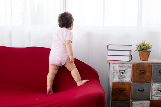 Portrait Asian toddler girl infant climbing red sofa at home looking to window, happy healthy cute baby kid soft skin waiting for mother or playing alone, Children's learning and child development.
