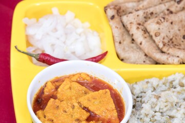Very Close-Up view of Gujarati Kathiyawadi Spicy Dhokali Sabji, khichdi, Roti, Onion Pieces, Red Chilli, on yellow square dish prominently, set against a red background