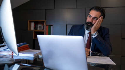 A middle-aged, dark-haired businessman in a suit is working at his desk in the office with his computer, looking bored