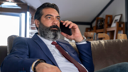 A middle-aged, dark-haired businessman in a suit is sitting on a leather couch in his office, having a tense business meeting on his phone