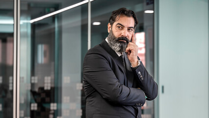 A middle-aged, businessman is in an empty office with a blurred background, shown in a close-up shot, resting his hand on his cheek and making facial expressions that indicate he is thinking