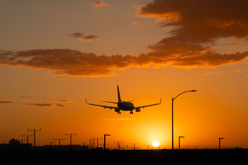 Airplane landing at Los Angeles International airport (LAX) with the sun setting. Orange sky and orange colored clouds. Airport infrastructure visible as dark silhouettes.	