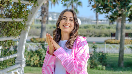Fototapeta premium A young brunette businesswoman in a tree-filled park on a spring day, looking at the camera while clapping her hands