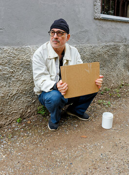 Elderly man with a beard, wearing a beret and glasses, sitting on the pavement holding a cardboard sign next to a white cup to collect money, Italy