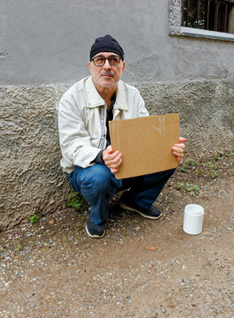 Middle-aged man squatting on the sidewalk holding a cardboard sign with a white container to collect money placed beside him, Italy