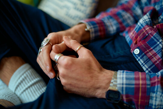 Close-up of a man with clasped hands wearing silver rings, checkered shirt, white socks and jeans.
