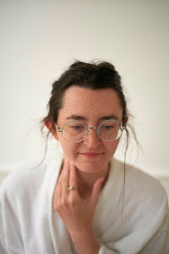 Close-up of a woman with glasses and a thoughtful expression wearing a white robe touching her neck gently against a neutral background.
