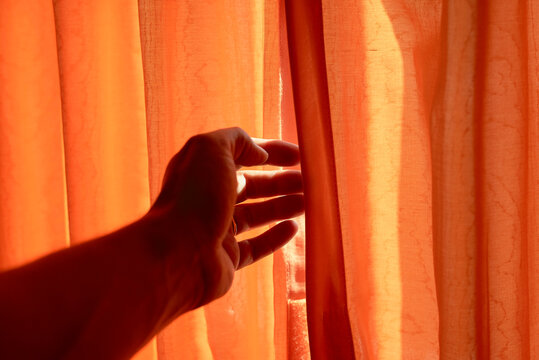 A hand is parting an orange curtain in a cargo ship cabin to let in sunlight.