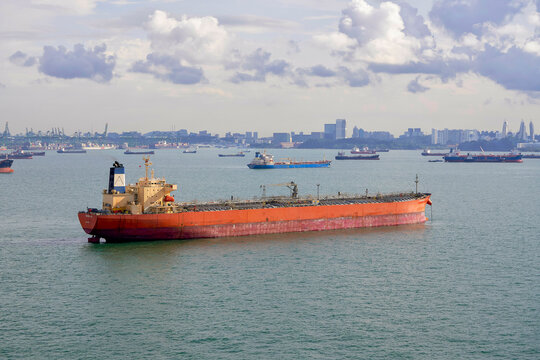 Cargo ship sailing on a vast sea with a fleet of vessels and a distant city skyline under a cloudy sky, Singapore