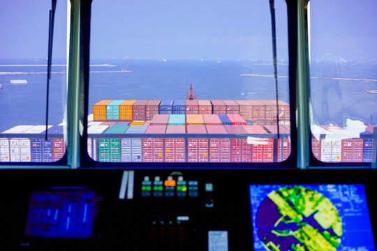 View from a ship's bridge with a cargo ship's container deck and ocean horizon in the background.