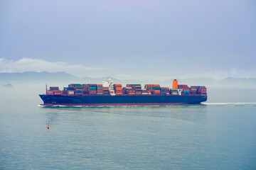 A cargo ship laden with containers sails across a misty sea with mountains faintly visible in the background, Busan, Korea