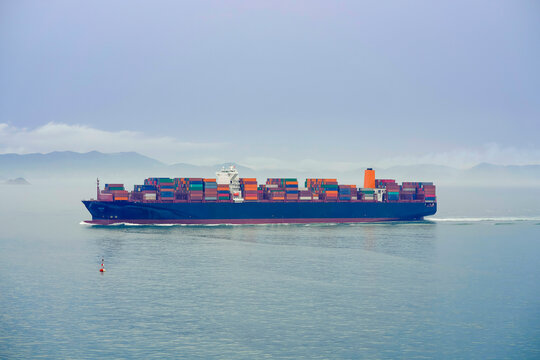 A cargo ship laden with containers sails across a misty sea with mountains faintly visible in the background, Busan, Korea
