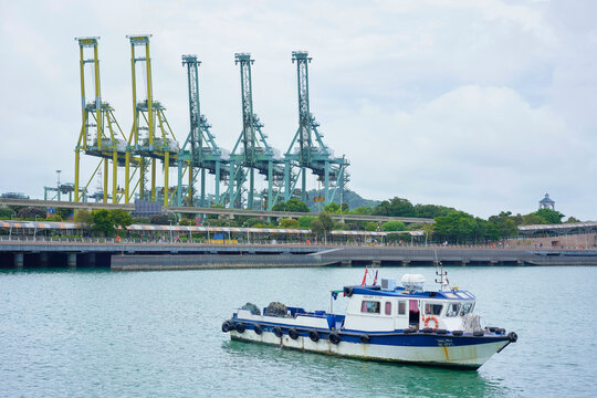 A tugboat sails in front of towering container cranes at a busy shipping port under a cloudy sky, Singapore