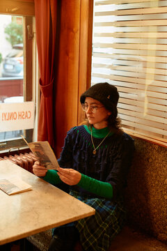A pensive person sits at a cafe table by the window, reading a postcard in a cozy, warmly lit interior.