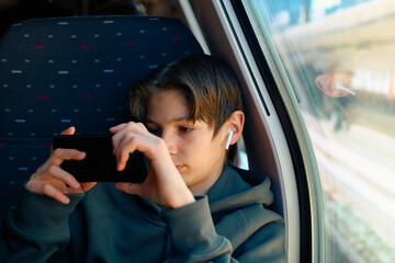 A teenager wearing a hoodie and wireless earbuds focuses intently on his smartphone while sitting by a train window.