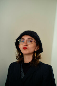 Stylish young woman with round glasses and black beret pouting her red lips against a light background.