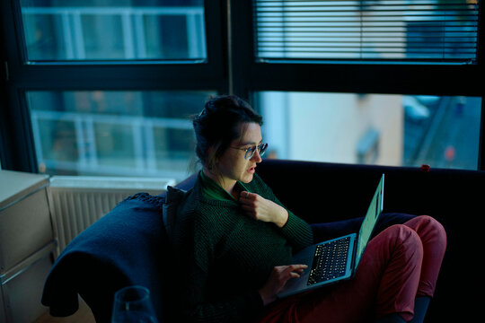 Woman in glasses using a laptop while sitting on the floor by a window at dusk.