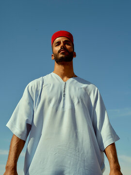 Confident man standing against a clear blue sky wearing a white tunic and red headband, Morocco