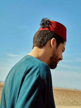 A man in a red fez and blue shirt gazes downward against a clear sky and arid background.