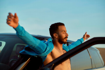 Man in a blue jacket leaning out of a car window with arms stretched against a clear sky at dusk, Morocco