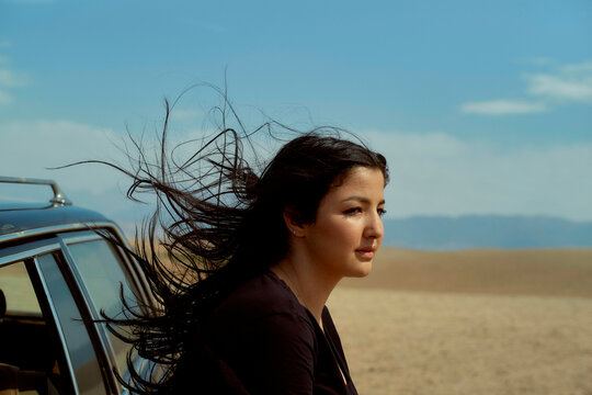 Woman with flowing hair looking into the distance next to her car in a desert setting, Morocco