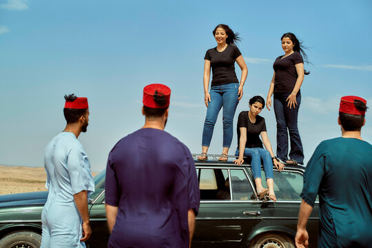Three women stand on top of a car with three men in traditional headwear facing them in a desert setting, Morocco