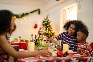 A joyful family gathers around the dinner table celebrating Christmas. The father, dressed in a Santa hat, gives a gift to the child, while everyone smiles with warmth and festive decorations.