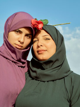 Two women in hijabs posing against a clear sky, one with a red flower adorned on her headscarf, Morocco