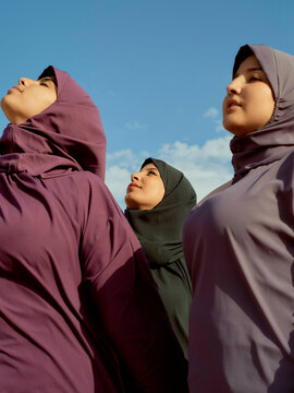 Three women in hijabs stand confidently against a blue sky, gazing upwards with poised expressions, Morocco