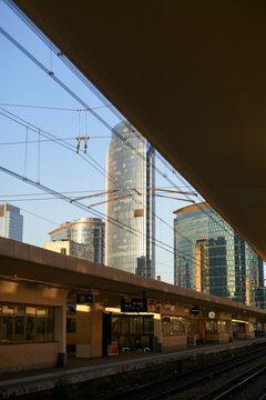 Skyscrapers tower over a train station platform during sunset with the sun casting a warm glow on the scene.
