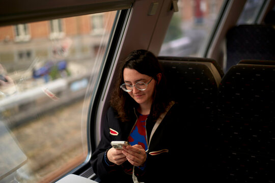 A woman smiles while looking at her smartphone in a train compartment, seated by the window with soft natural light highlighting her face, Belgium
