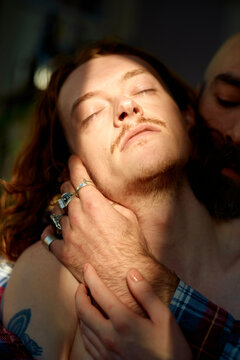 Young man with closed eyes receives a gentle caress on the cheek by his partner, bathed in soft sunlight, evoking a sense of intimacy and tenderness.