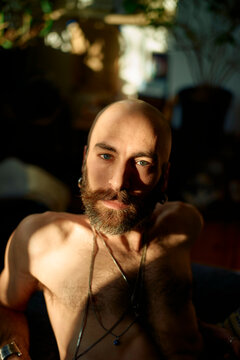 Shirtless man with a beard and necklace sitting in natural sunlight.