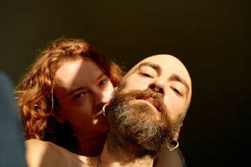 Intimate moment of a gay couple with a young bearded man being embraced, caressed, and kisses from behind by his tattooed partner, bathed in warm sunlight.