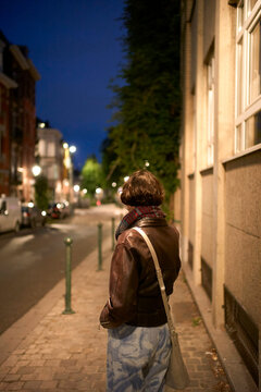 A young woman in a leather jacket and scarf stands on a city street at night, facing away from the camera.