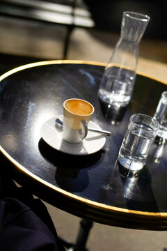 An empty white espresso cup with coffee stains on a saucer with a coffee spoon, and a glass of water on a round black table.